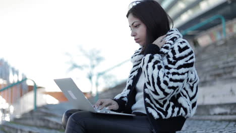 Concentrated-young-woman-sitting-outdoor-with-laptop-on-knees